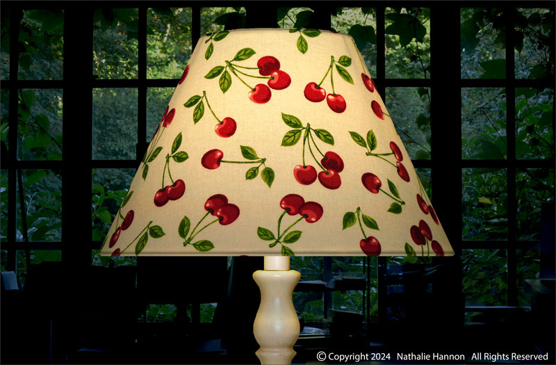 Éclairage d'un abat-jour déco de forme conique sur pied de lampe en tissu coton à motif cerises et feuilles.